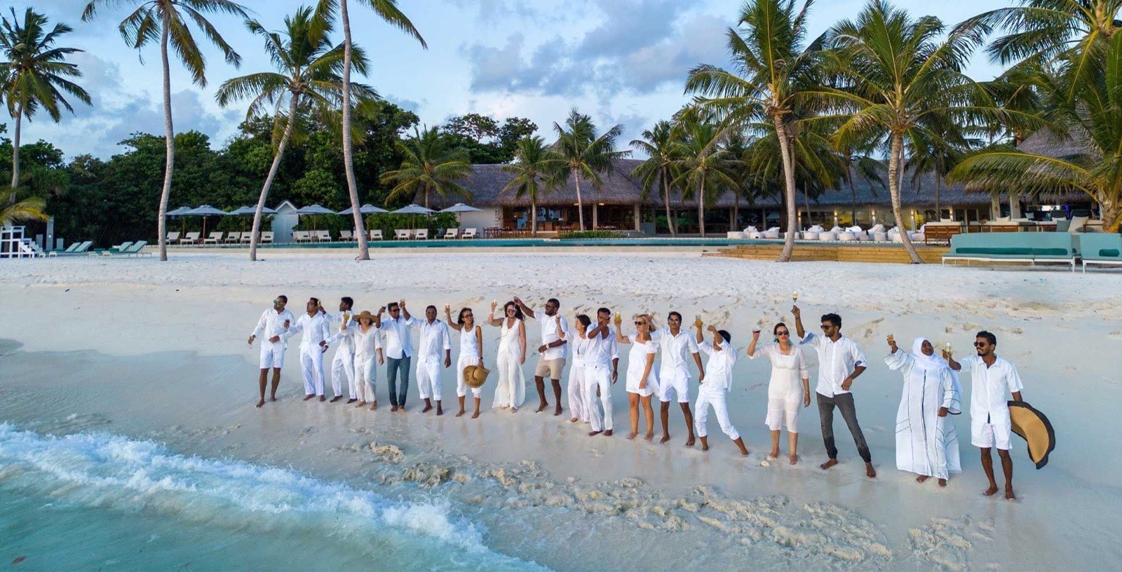 A large group dressed in white standing along the water's edge at Ifuru Island, raising glasses together, with the resort's pool and pavilions behind them.